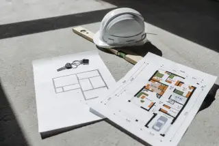 Illustration: A hard hat, level, keys, and floor plan blueprints resting on a sunlit concrete surface.