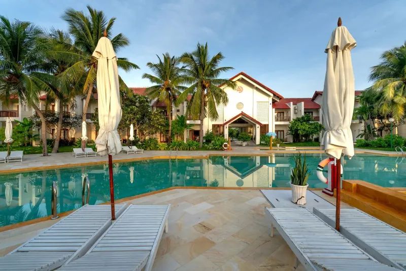Illustration: Sun loungers and parasols by an empty swimming pool in front of a tropical resort building.